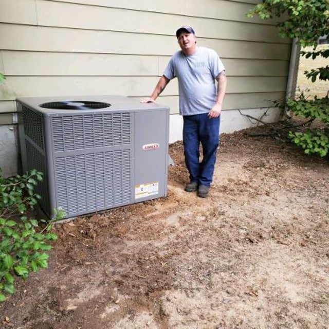 Customer leaning against new Lennox Heating and Air Conditioning System outside of his house on slab of concrete surrounded by dirt.