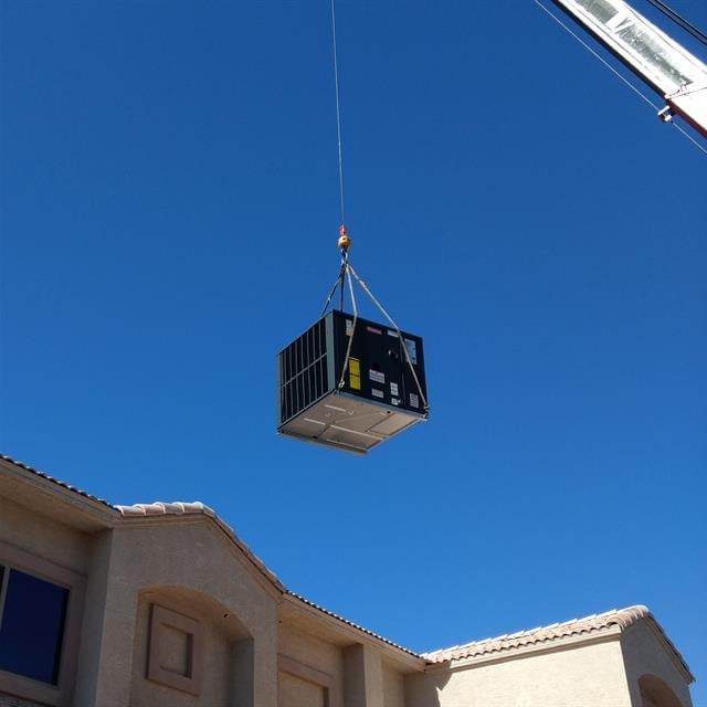 An AC unit suspended in the air by a crane, about to be installed on a roof.