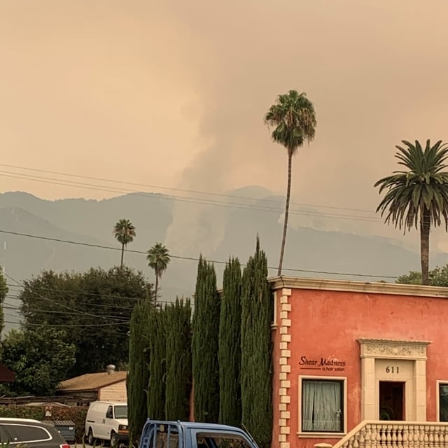 A fire in the mountains near Pasadena, with a terracotta building and palm and cypress trees in the foreground