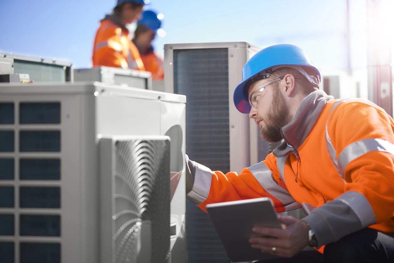 An air conditioning technician checks the measurements on a unit with an orange vest, hard hat, and clipboard in hand.