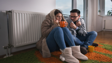 A man and woman sit next to their radiator while wrapped in blankets, indicating that their home’s heating system is broken. | Aire Serv of North Central Arizona