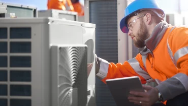 An air conditioning technician checks the measurements on a unit with an orange vest, hard hat, and clipboard in hand.