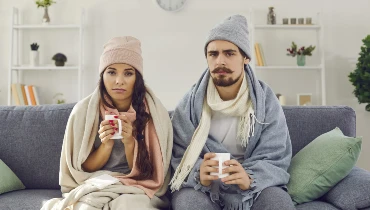 Upset young couple sitting on the couch, bundled up and having problem with central heating