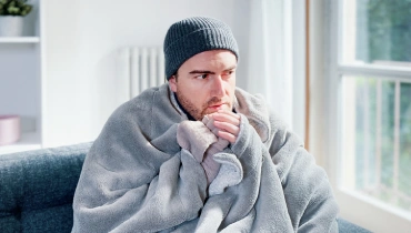 A man wearing a wool beanie and wrapped in a blanket sitting on a couch in a living room