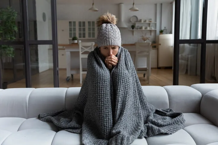  A woman wrapped in a blanket on her couch because her home’s heater is broken.