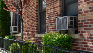 Outdoor window air conditioning units on an old New York City brick apartment building with green plants along a sidewalk | Aire Serv of Springfield, Illinois