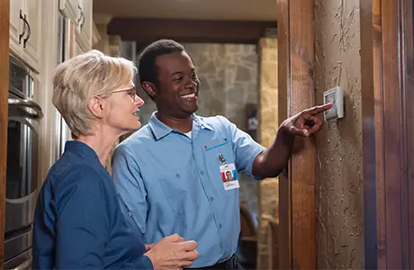 Male Aire Serv technician adjusting thermostat beside female customer in blue shirt.