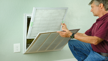 Man in wine-colored polo shirt and blue jeans replacing home air vent filter.