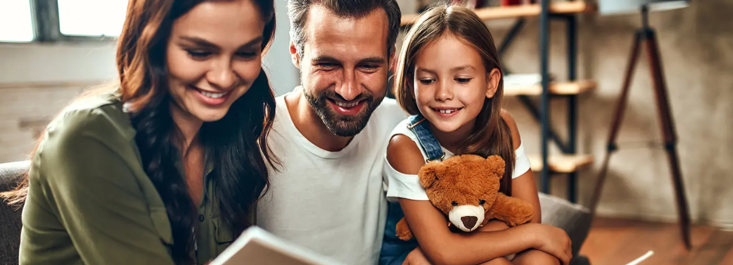 Smiling couple reading storybook to daughter holding teddy bear in living room interior.